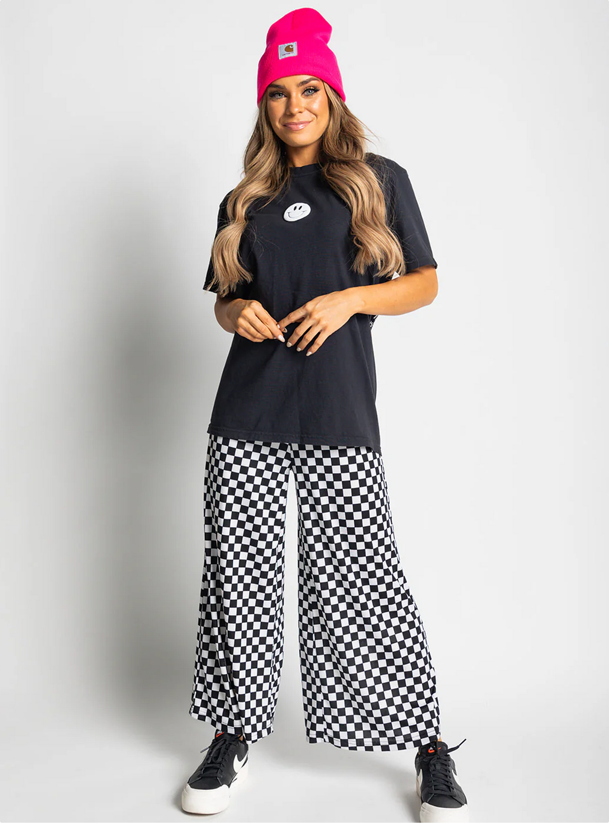 Woman wearing a black t-shirt, pink beanie, and checkered pants on a white background